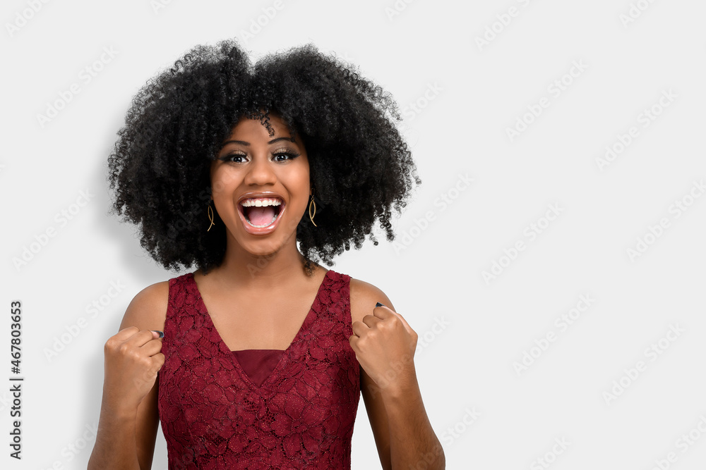 A cheerful and happy young girl with afro hair proves to be the winner, raises her clenched fists in celebration. A beautiful dark-skinned girl overflows with joy or happiness, is dressed in a red dre A cheerful and happy young girl with afro hair proves to be the winner, raises her clenched fists in celebration. A beautiful dark-skinned girl overflows with joy or happiness, is dressed in a red dre