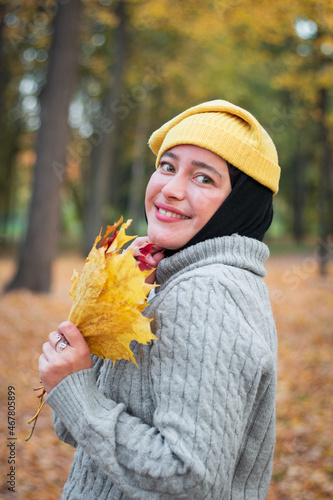 Young Muslim woman in a yellow hat with a gray sweater with  a yellow autumn leaf. Autumn Park. Femininity and a smile