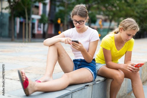 Teenager girls in shorts sitting in square with smartphones in hands.