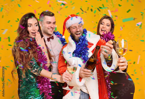 a group of friends is celebrating a Christmas party.A young man in a Santa Claus costume opens a bottle of champagne and has fun with friends in smart clothes against the background of flying confetti
