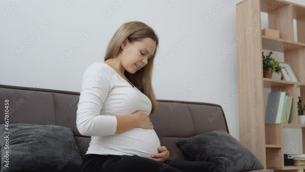 A pregnant fair-skinned woman is sitting on a cozy sofa and stroking her tummy A pregnant fair-skinned woman is sitting on a cozy sofa and stroking her tummy