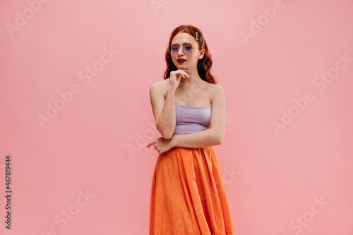 Slim caucasian young girl looks into camera holding chin with hand on pink background. Beauty with red flowing hair in round glasses, lilac top, orange skirt. Emotional reaction to happiness, concept