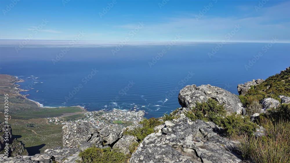 The blue Atlantic Ocean and azure sky are visible from the top of Table Mountain in Cape Town. An urban area on an uneven coastline. There are grey spotted boulders in the foreground. South Africa The blue Atlantic Ocean and azure sky are visible from the top of Table Mountain in Cape Town. An urban area on an uneven coastline. There are grey spotted boulders in the foreground. South Africa