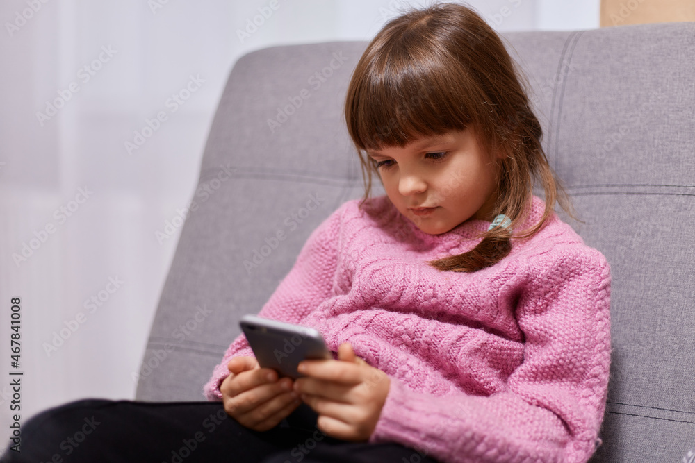 Portrait of dark haired little girl with pigtails wearing warm pink sweater, playing video game, sitting on sofa in living room, spending free time with gadget. Portrait of dark haired little girl with pigtails wearing warm pink sweater, playing video game, sitting on sofa in living room, spending free time with gadget.