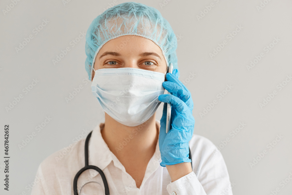 Smiling positive female doctor wearing medical uniform, cap and surgical mask talking phone with patient, looking at camera with optimistic facial expression. Smiling positive female doctor wearing medical uniform, cap and surgical mask talking phone with patient, looking at camera with optimistic facial expression.