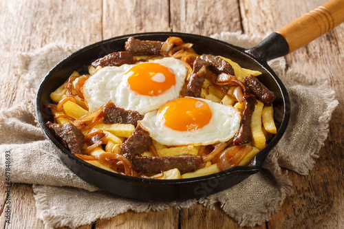 Chorrillana is a favorite Chilean dish of French fries loaded with beef, caramelized onions, and fried eggs close up in the pan on the table. Horizontal photo