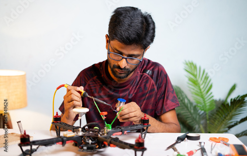 Young man or student making drone by soldering wires to baord at home - concept of drone repair and develoment, scientific experimenting or researching. photo