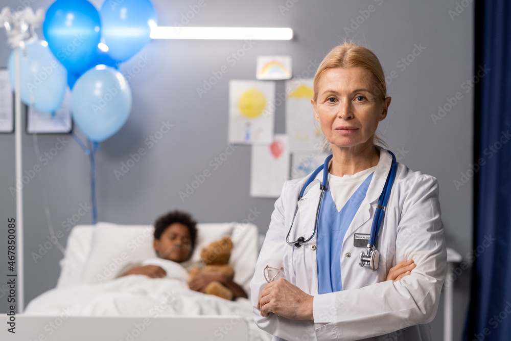 Portrait of mature pediatrician in white coat standing with her arms crossed and looking at camera with little patient in the background