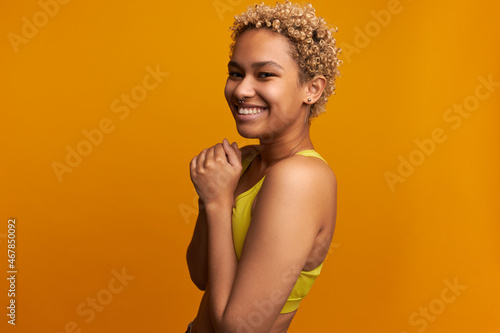 Horizontal indoor portrait of blonde dark-skinned young cute charismatic girl of 20s in perfect mood, isolated on orange studio wall, laughing standing half-turned to camera in yellow sport top