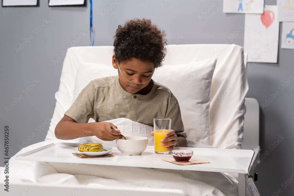 Sick little boy sitting in bed with tray and having breakfast while being treated at hospital Sick little boy sitting in bed with tray and having breakfast while being treated at hospital