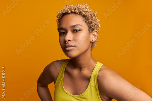 Horizontal close-up of beautiful charming youthful ethnicity girl with blonde short curls, wearing nose jewelery, looking at camera with confident face, holding head high. People and personality