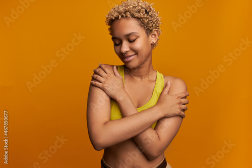 Cute African female with short little blonde poodles posing with arms around her shoulders, enjoying beauty of her body dressed in yellow sport top, wearing nose ring, smiling with embarrassment