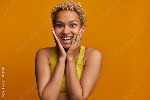 Happy excited stylish blondie with dark skin, short curls and pierced nose, holding cheeks with hands, hearing perfect news, winning lottery, surprised with best results of sports competition