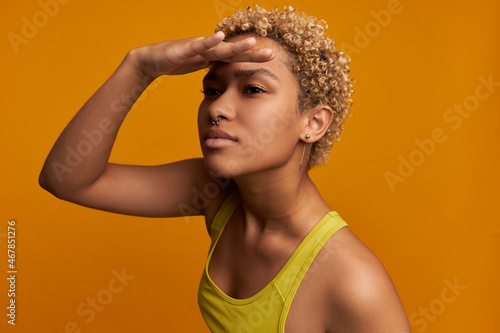 Body language concept. Mixed-race blondie looking far in distance covering eyes from sun with palm over her forehead, standing against orange wall at studio, wearing yellow, nose and ears jewelery