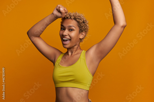 Horizontal picture of beautiful attractive fitness girl jumping with happy excited face expression, putting hands up, rejoicing, dressed in yellow isolated on orange. Human emotions and feelings