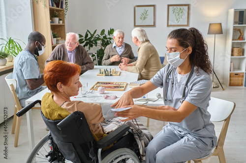 Nurse measuring the pressure to senior woman who using wheelchair in the nursing home with other senior people playing chess in the background