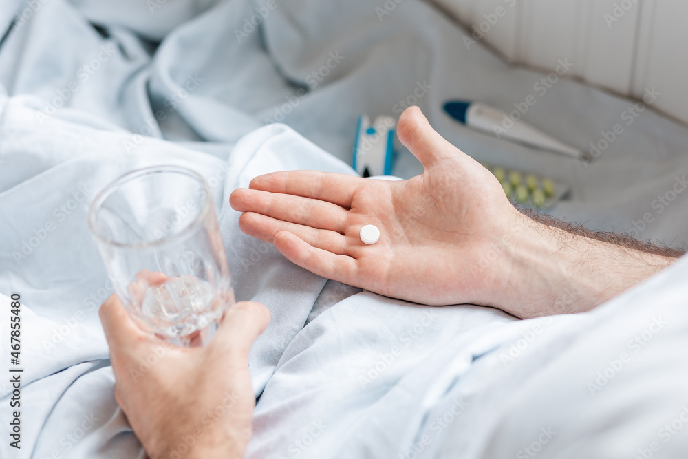 A man is lying in bed with a glass of water and holding a pill in the palm of his hand. Close up. Top view A man is lying in bed with a glass of water and holding a pill in the palm of his hand. Close up. Top view