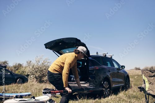 Windsurfer and camper packing and unpacking from a car's roof rack in nature.