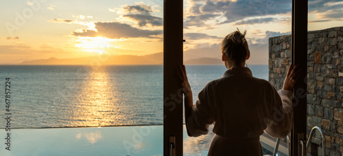 woman in bathrobe standing between sliding glass doors and enjoying sunrise over the sea in luxury villa © ronstik