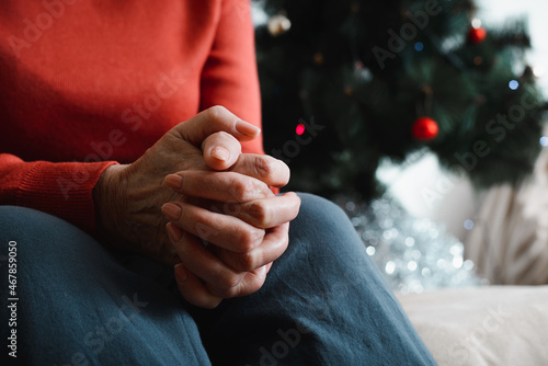 Lonely senior woman sitting at home in Christmas celebration. Close-up of an elderly woman's hand against background of decorated Christmas tree. Loneliness, sad holiday concept
