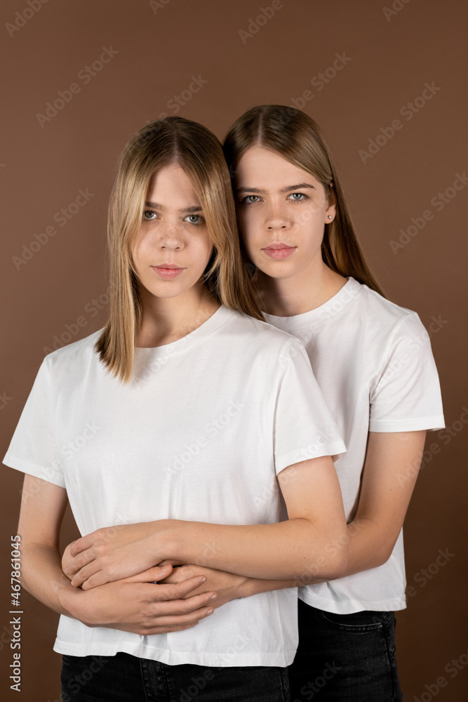 One of affectionate twin girls in white t-shirts embracing her sister while both standing against brown background One of affectionate twin girls in white t-shirts embracing her sister while both standing against brown background