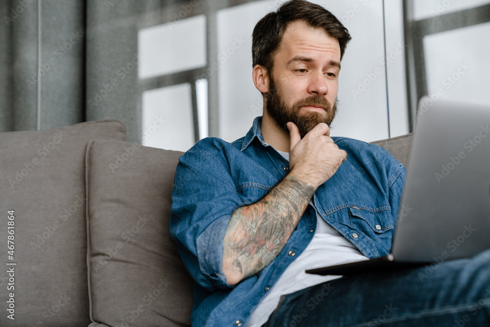 Bearded european man working with laptop while sitting on sofa at home