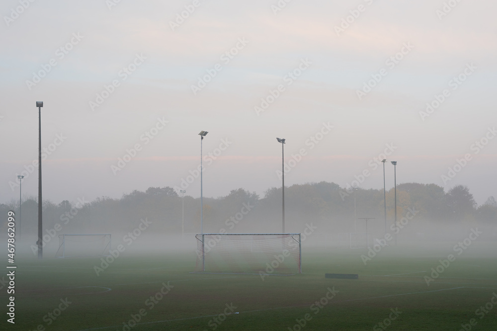 Empty and deserted soccer field in suburban area with goals and flood light poles on grey overcast day with fog and mist - obrazy, fototapety, plakaty 