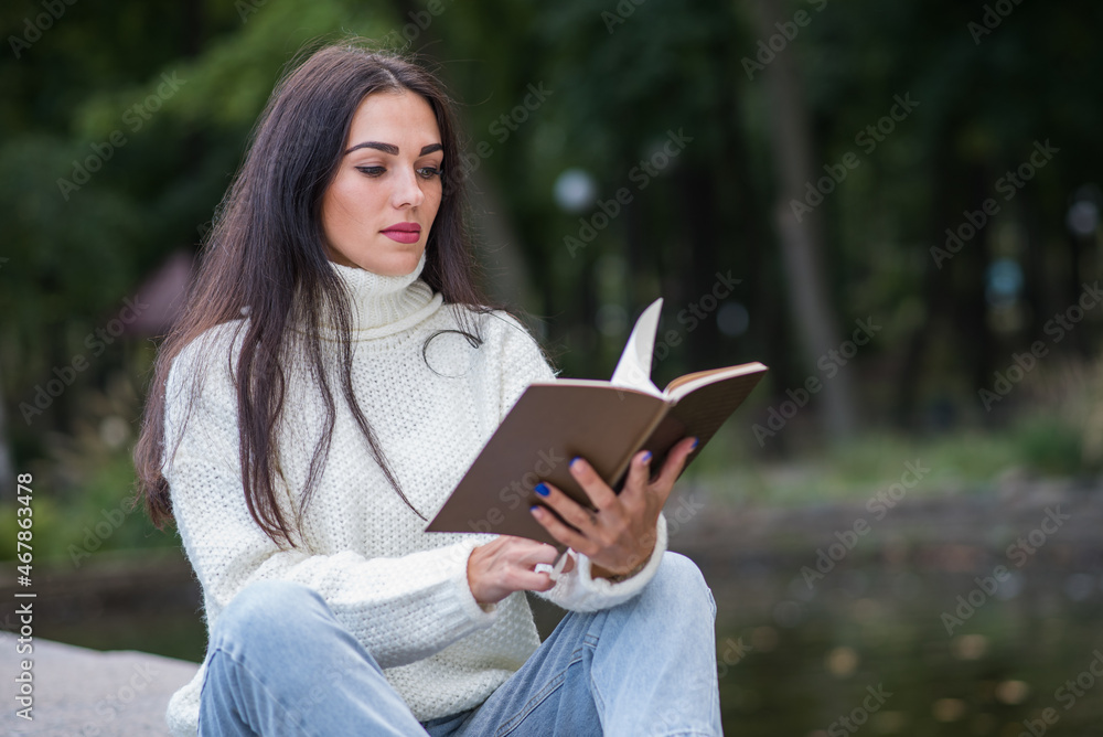 Obraz premium A young and attractive Caucasian brunette girl sitting and reading a book in a park near the river. The student is studying. The concept of education and knowledge.