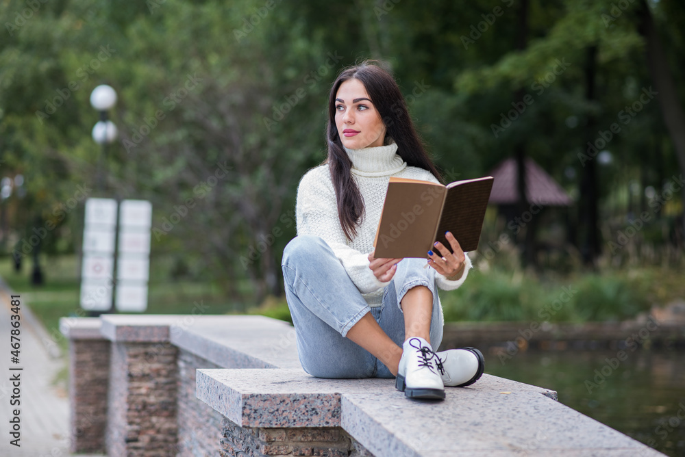 Obraz premium A young and attractive Caucasian brunette girl sitting and reading a book in a park near the river. The student is studying. The concept of education and knowledge.