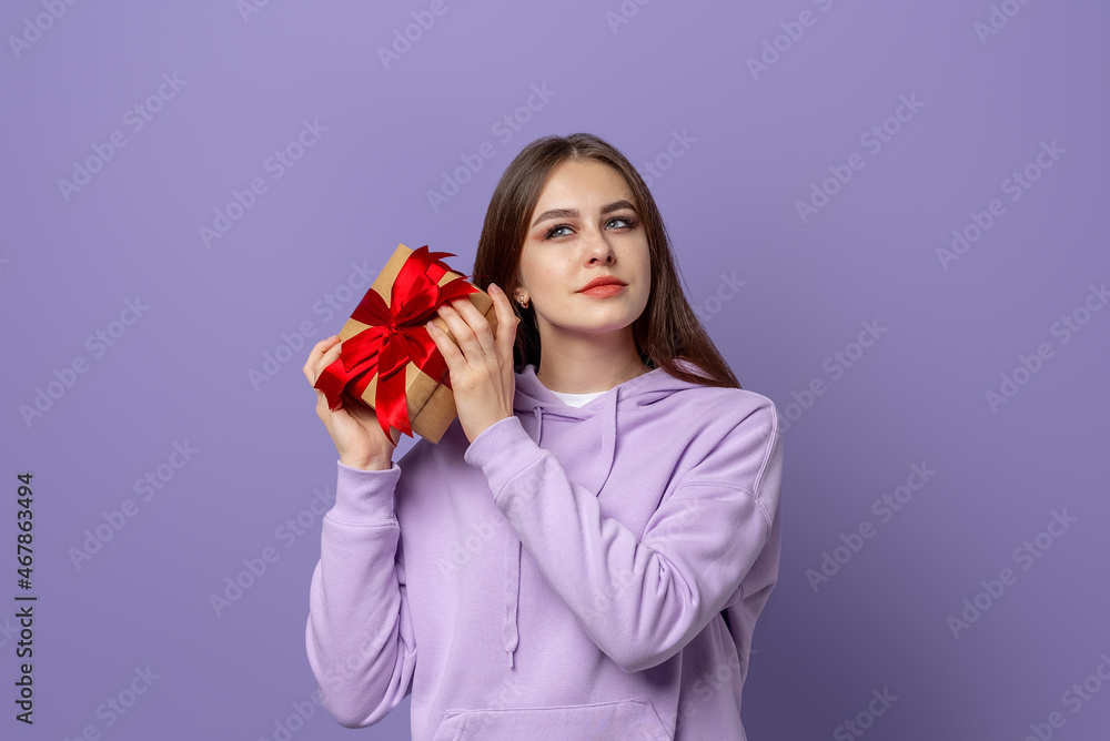 Happy young brunette woman dressed casual hoodie, standing isolated over purple background, holding a gift box Happy young brunette woman dressed casual hoodie, standing isolated over purple background, holding a gift box