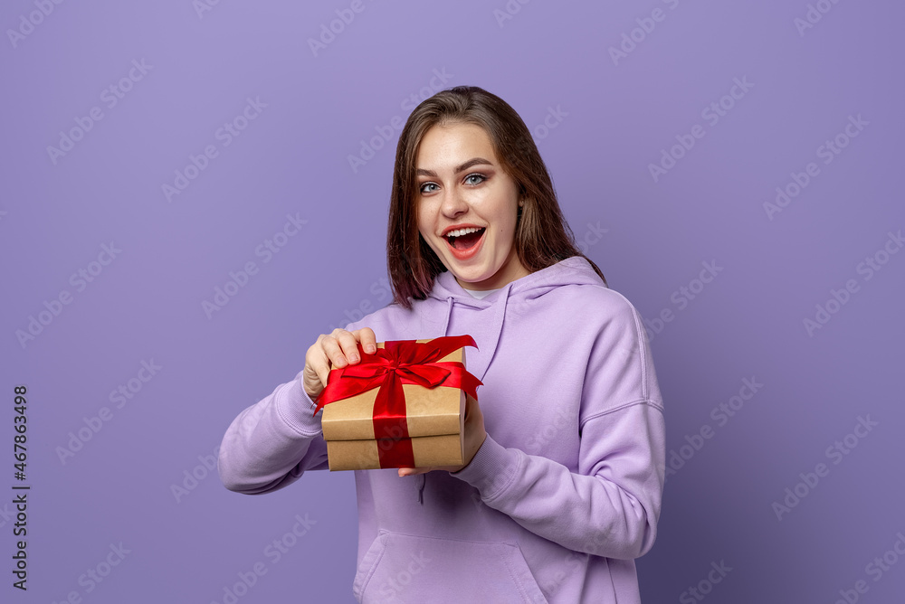 Happy amazed brunet woman in purple hoodie opening a present, standing over purple background. New Year Women's Day birthday holiday concept Happy amazed brunet woman in purple hoodie opening a present, standing over purple background. New Year Women's Day birthday holiday concept