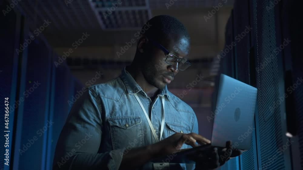Engineer network specialist works in server room. Afro-American guy in denim shirt spbas runs system diagnostics at laptop screen illumination in dark corridor closeup Engineer network specialist works in server room. Afro-American guy in denim shirt spbas runs system diagnostics at laptop screen illumination in dark corridor closeup