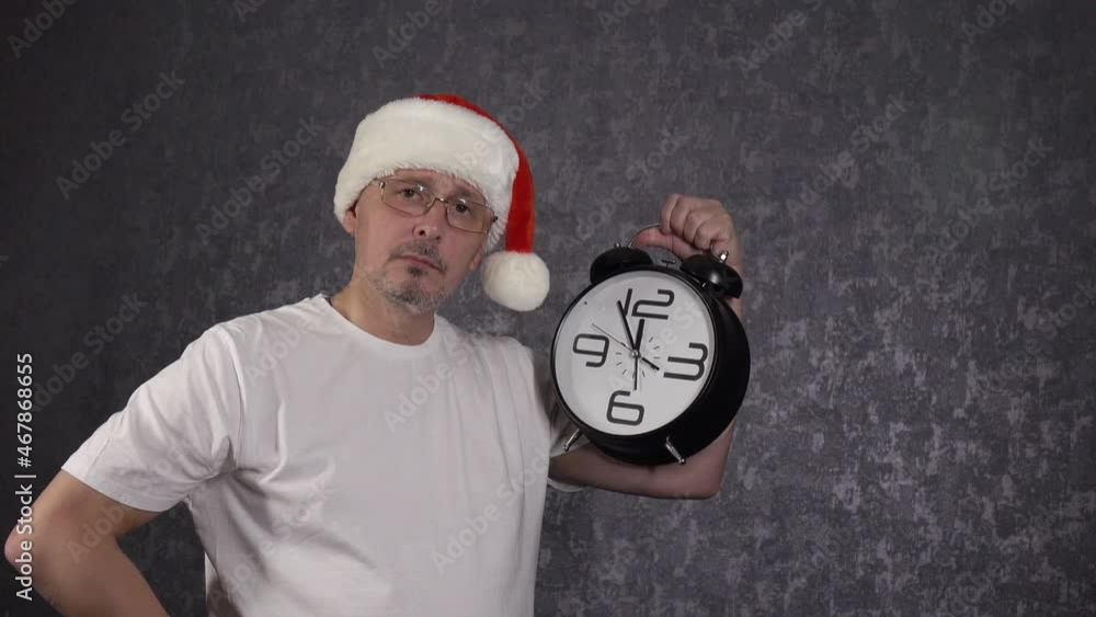 A man shows his watch and is happy to finish work before the New Year, dressed in a T-shirt and a Santa Claus hat. A man shows his watch and is happy to finish work before the New Year, dressed in a T-shirt and a Santa Claus hat.