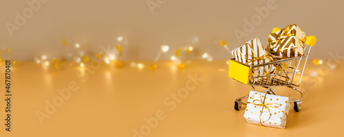 Online christmas shopping and sale. Shopping cart with gift boxes on golden background with lights. Supermarket trolley full presents. Banner with copy space