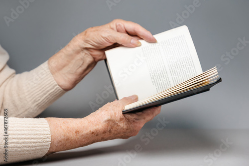 Wrinkled hands of a senior woman are holding a book. Gray background. The concept of education and leisure