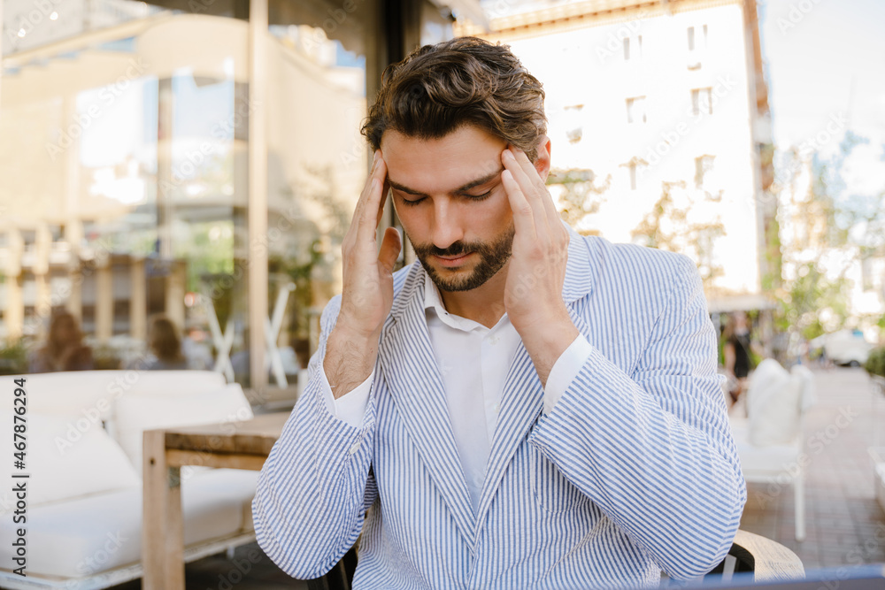 Young man with headache rubbing his temples while sitting at cafe Young man with headache rubbing his temples while sitting at cafe