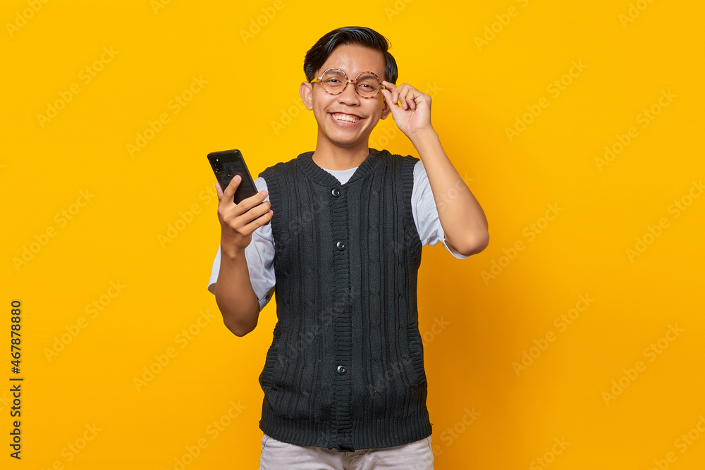 Smiling Asian man touching glasses and holding mobile phone in one hand over yellow background Smiling Asian man touching glasses and holding mobile phone in one hand over yellow background