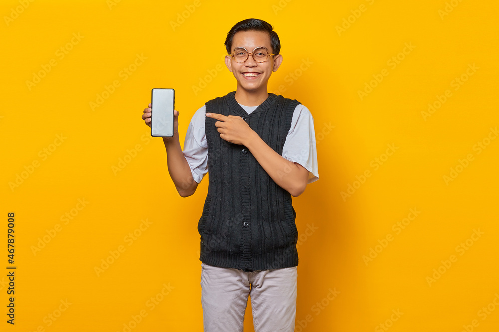 Smiling handsome young man pointing to blank smartphone screen on yellow background Smiling handsome young man pointing to blank smartphone screen on yellow background