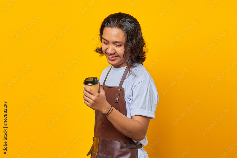 Portrait of handsome man wearing apron looking at cup of coffee being held isolated on yellow background Portrait of handsome man wearing apron looking at cup of coffee being held isolated on yellow background