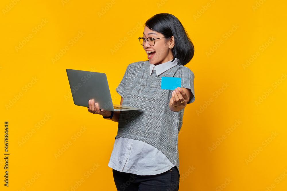 Cheerful asian woman showing credit card and looking at laptop over yellow background Cheerful asian woman showing credit card and looking at laptop over yellow background