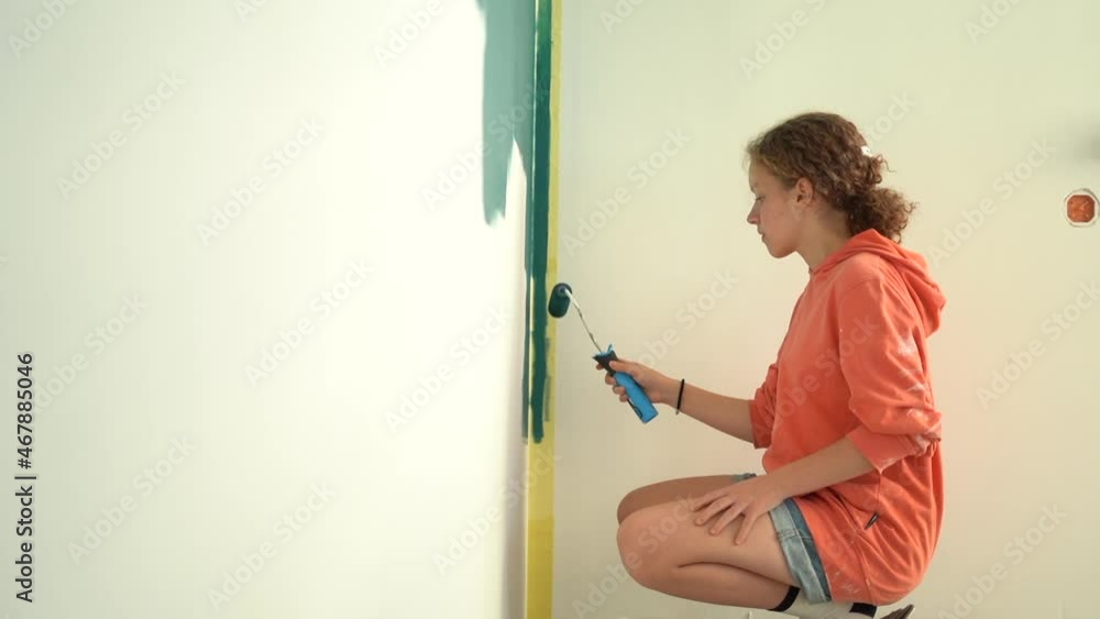 Teenage girl with red curly hair paints the wall blue with a roller in her own room. Renovation and creativity concept. The girl sits on his haunches, crouched near the wall with a paint tool Teenage girl with red curly hair paints the wall blue with a roller in her own room. Renovation and creativity concept. The girl sits on his haunches, crouched near the wall with a paint tool
