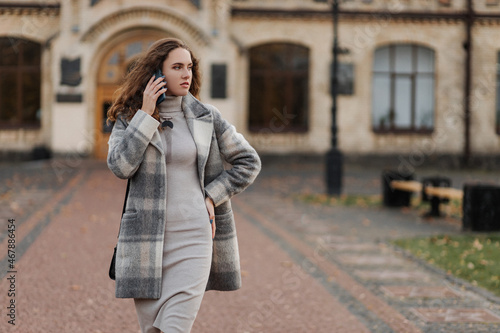 Young girl student in a gray coat talking by phone of the university background 