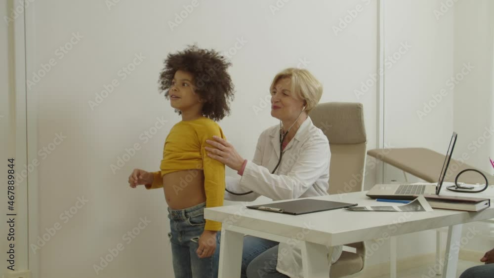 Caring beautiful professional middle aged woman physician in lab coat auscultating lung of lovely elementary age african american child with stethoscope, making diagnosis for sick patient at hospital. Caring beautiful professional middle aged woman physician in lab coat auscultating lung of lovely elementary age african american child with stethoscope, making diagnosis for sick patient at hospital.