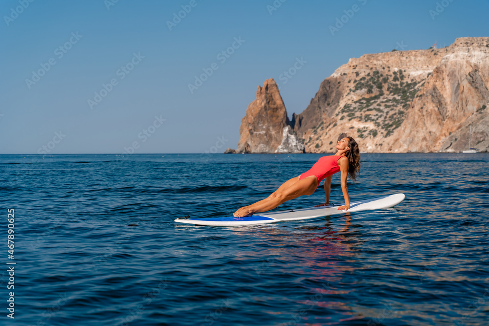 Sporty girl on a surfboard in the sea on a sunny summer day. In a red swimsuit, she sits in the splits on the sap. Summer entertainment on Stortom by the sea