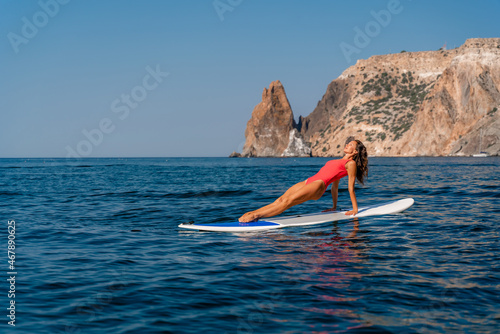 Sporty girl on a surfboard in the sea on a sunny summer day. In a red swimsuit, she sits in the splits on the sap. Summer entertainment on Stortom by the sea