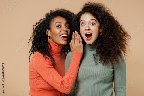 Two amazed young curly black women friends 20s wearing casual shirts clothes whispering gossip and tells secret behind her hand sharing news isolated on plain pastel beige background studio portrait.