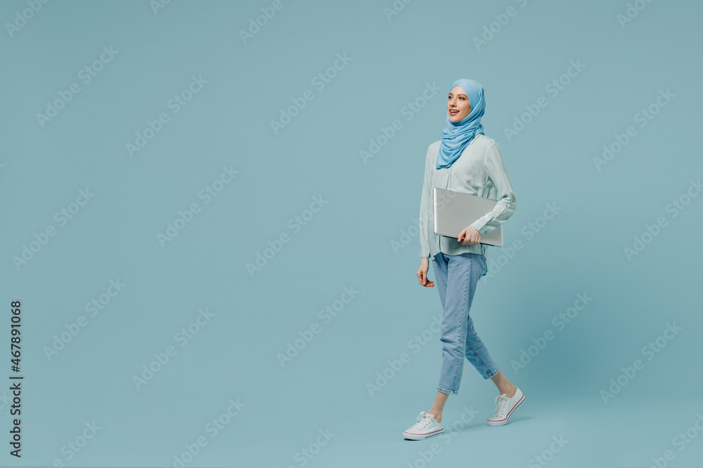 Full body side view young arabian asian muslim woman in abaya hijab hold closed laptop pc computer walking isolated on plain blue background studio. People uae middle eastern islam religious concept Full body side view young arabian asian muslim woman in abaya hijab hold closed laptop pc computer walking isolated on plain blue background studio. People uae middle eastern islam religious concept