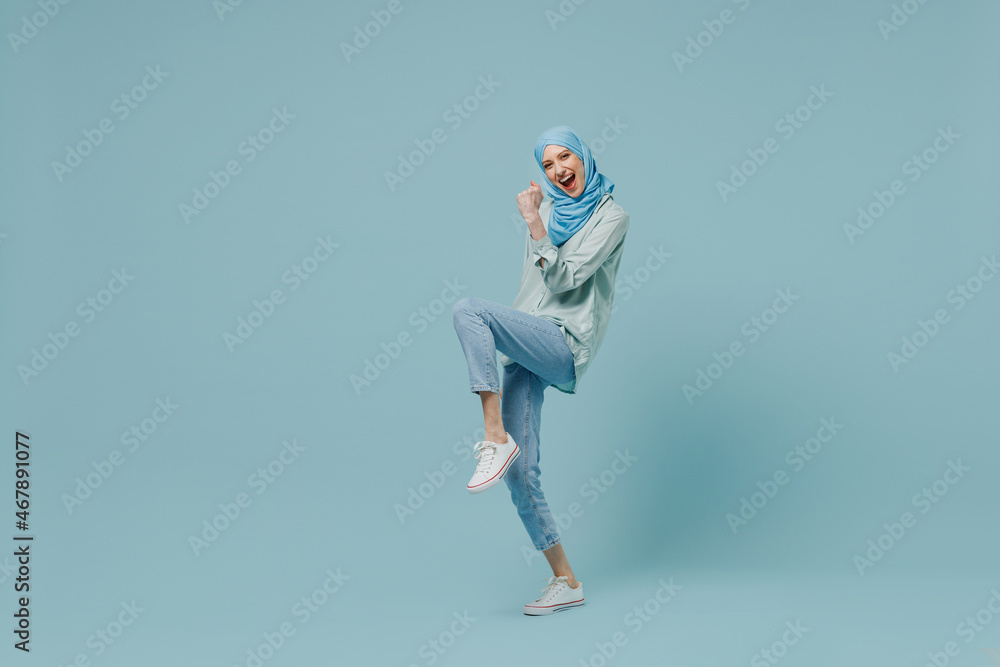 Full body overjoyed young arabian asian muslim woman in abaya hijab do winner gesture clench fist isolated on plain blue background studio portrait. People uae middle eastern islam religious concept. Full body overjoyed young arabian asian muslim woman in abaya hijab do winner gesture clench fist isolated on plain blue background studio portrait. People uae middle eastern islam religious concept.