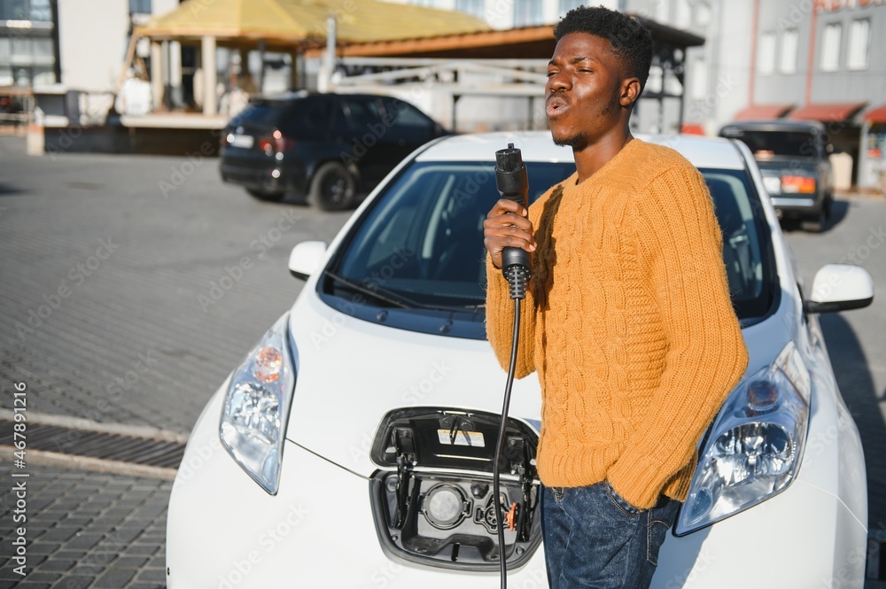 african man holding charge cable in on hand standing near luxury electric car.