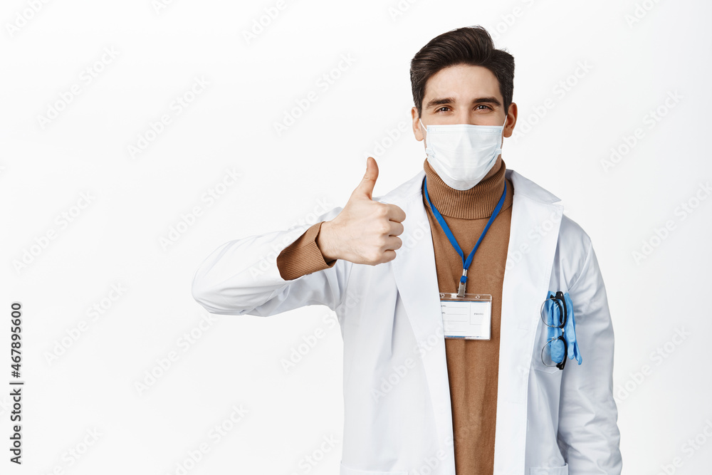 Covid-19 and pandemic. Smiling doctor in medical face mask, showing thumb up, standing in white robe over studio background Covid-19 and pandemic. Smiling doctor in medical face mask, showing thumb up, standing in white robe over studio background